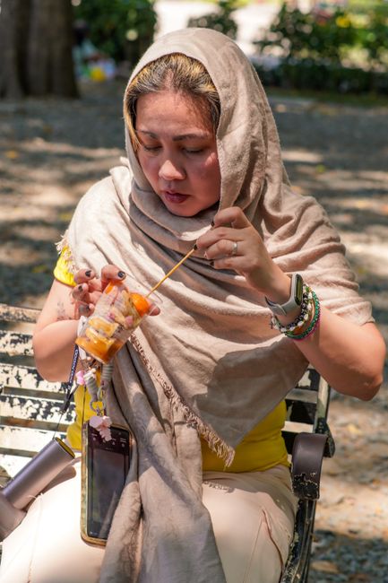 A woman in traditional attire enjoying a fruit snack on a park bench during a sunny day.