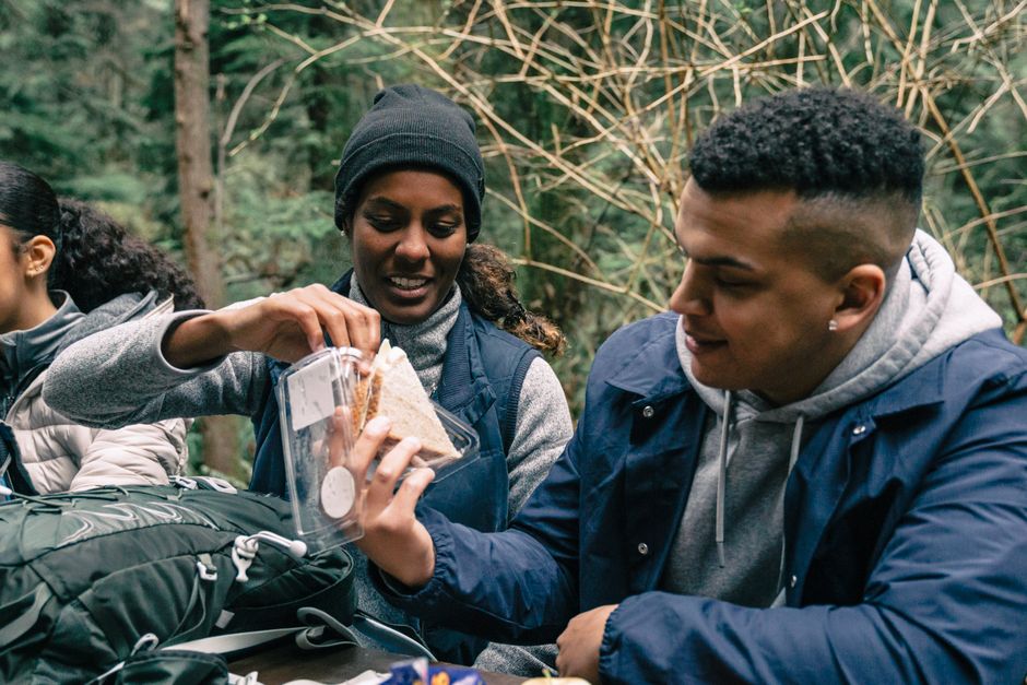 A group of friends enjoys a snack break in the forest, sharing sandwiches during a hiking adventure.
