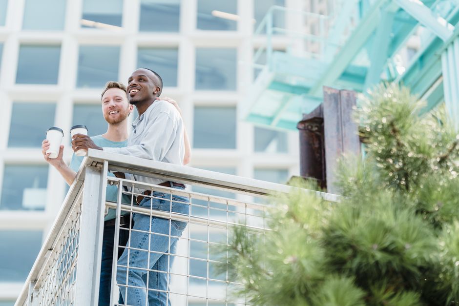 Interracial couple smiling and bonding over coffee on a sunny balcony in an urban setting.
