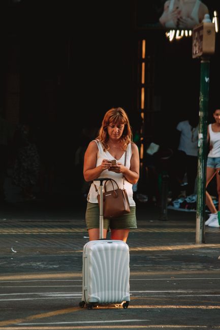 A woman standing with her luggage, checking her phone on a city street during the day.