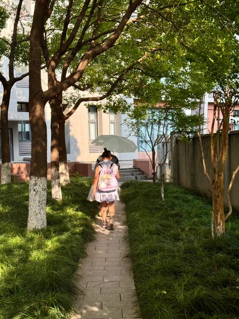 A woman with an umbrella walks down a tree-lined path in a sunny garden.