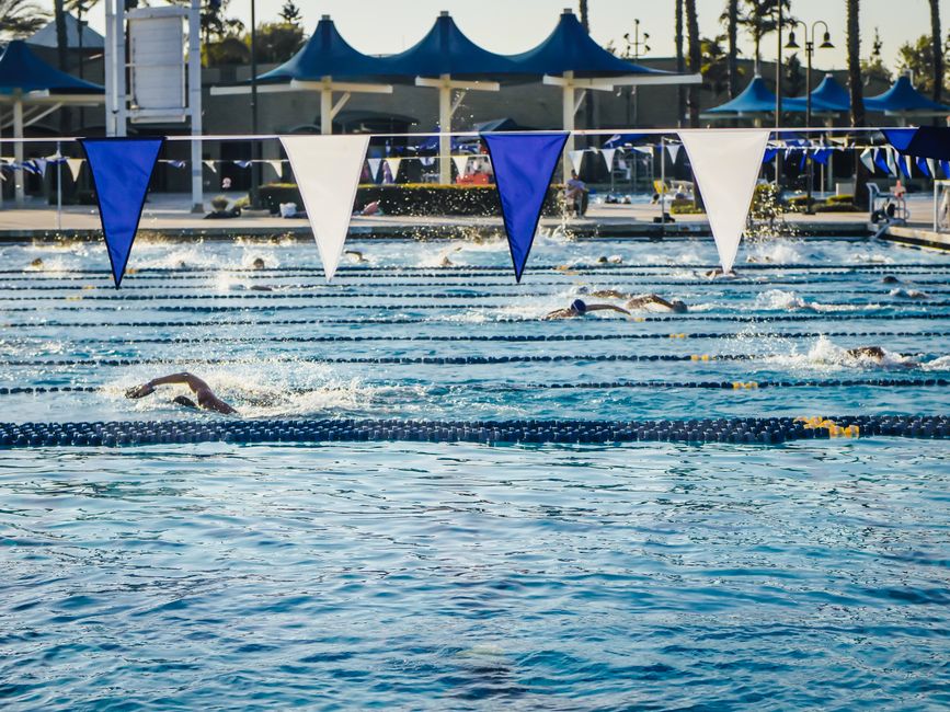Swimmers competing in an outdoor pool under blue and white banners on a sunny day.