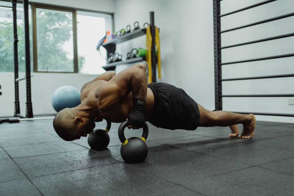 A shirtless man with a muscular build exercises in a gym, performing push-ups using kettlebells.