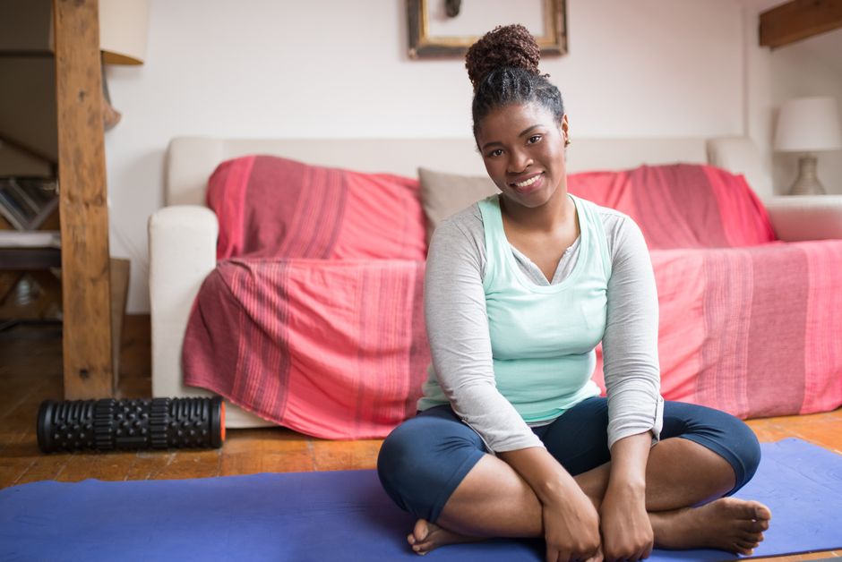 Smiling woman sitting on a yoga mat indoors, promoting a healthy lifestyle in a cozy home environment.