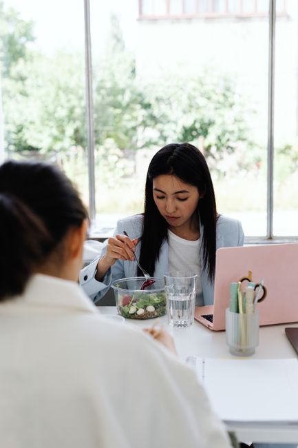 A young woman sits at her desk, enjoying a healthy salad lunch beside her laptop.