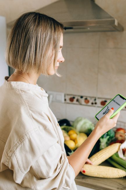 Woman with short hair using smartphone in kitchen, surrounded by fresh vegetables.