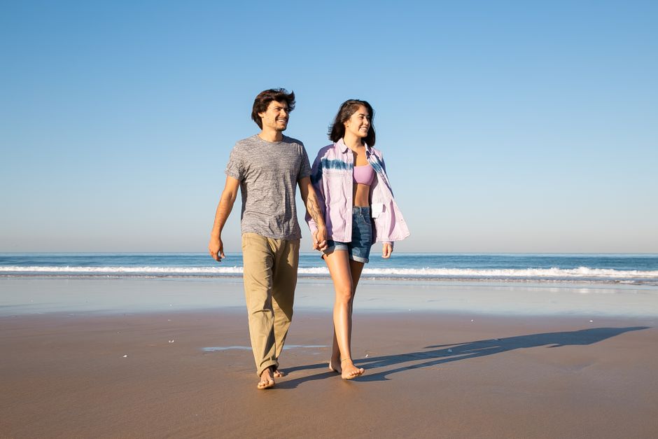 Smiling couple walking barefoot on a sunny beach, enjoying a romantic day outdoors.
