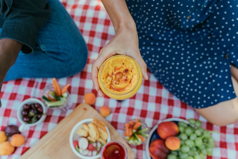 Top view of a colorful picnic spread featuring hummus, fruits, and vegetables on a checkered blanket.