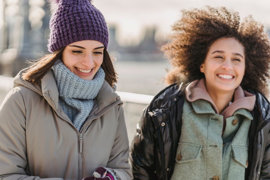 Happy young multiracial female friends in warm clothes strolling together on modern city promenade on sunny cold day