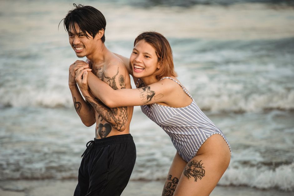 Joyful couple with tattoos enjoying a playful moment by the beach in summer.