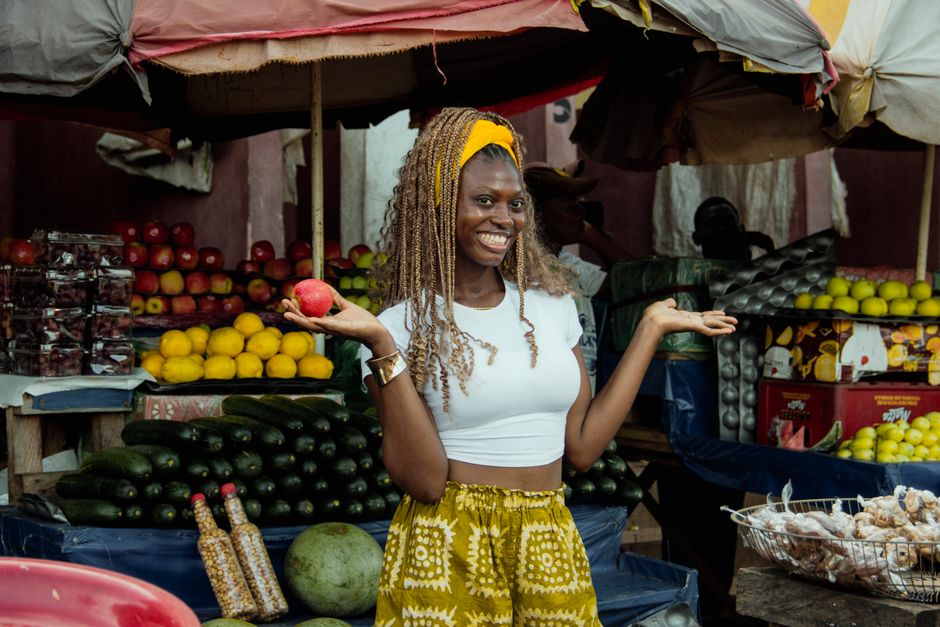Woman smiling while holding fruit in a colorful market setting, surrounded by fresh produce.