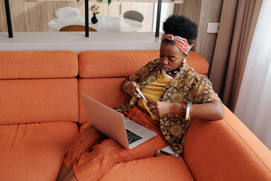 A worried woman sitting on an orange sofa checking her temperature with a thermometer, while a laptop sits open beside her.