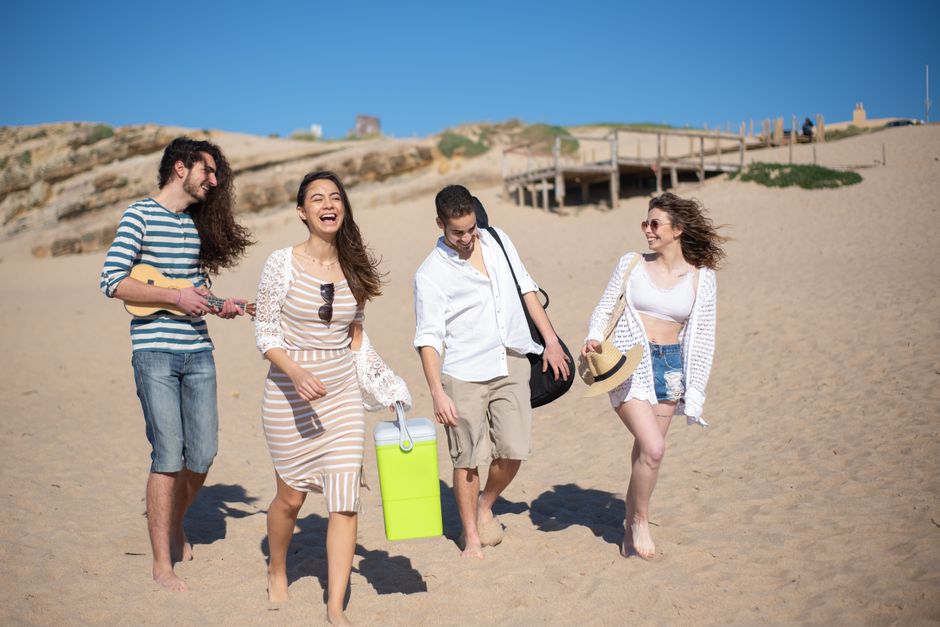 A group of friends laughing and walking barefoot on a sunny beach day in Portugal.