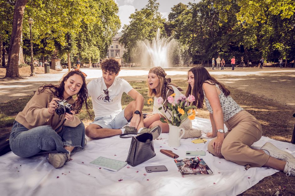 Happy group of friends having a picnic by a fountain in a Brussels park, enjoying summer outdoors.