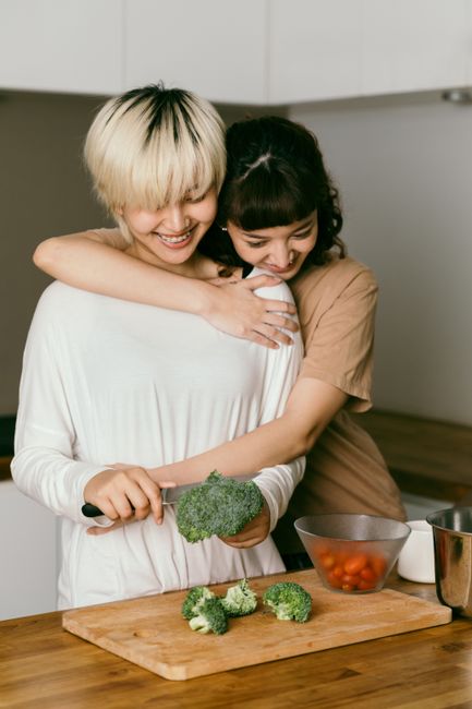 Happy loving Asian female couple in casual clothing cutting vegetables and cooking healthy dinner while bonding and smiling in sunny kitchen