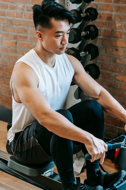 From above of stylish ethnic male in activewear exercising with rowing machine in sport club with dumbbells