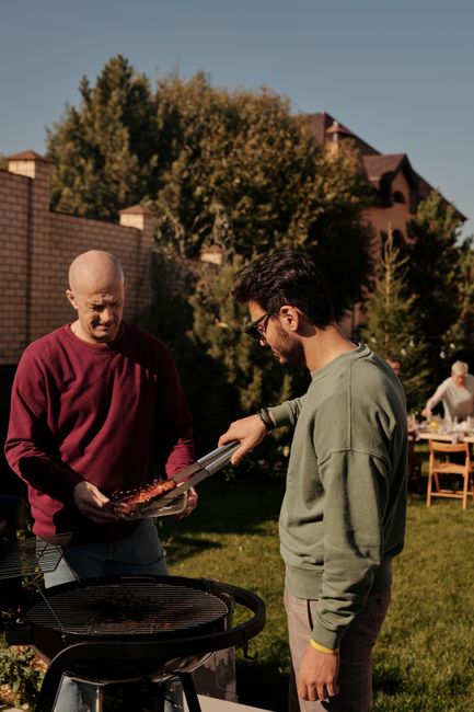 Two men grilling meat in a sunlit backyard, enjoying a barbecue with friends.