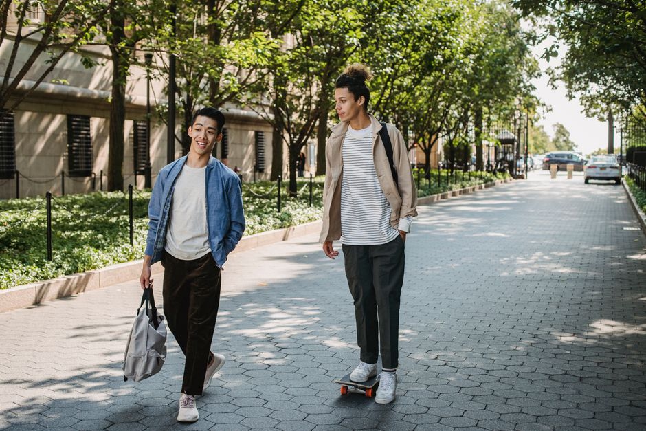 Full body positive young multiracial male friends wearing casual outfits chatting while riding skateboard and strolling together along sunny paved walkway in modern city