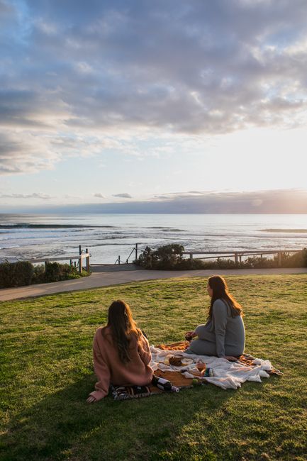 Back view of faceless female friends sitting on plaid with food and drink while enjoying view of sea during picnic