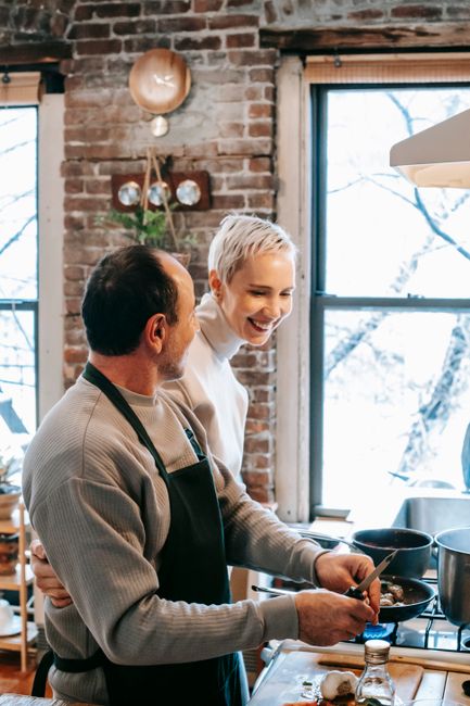 Smiling adult couple cooking lunch together near counter and ingredients near stove with pans near window in daytime in kitchen at home