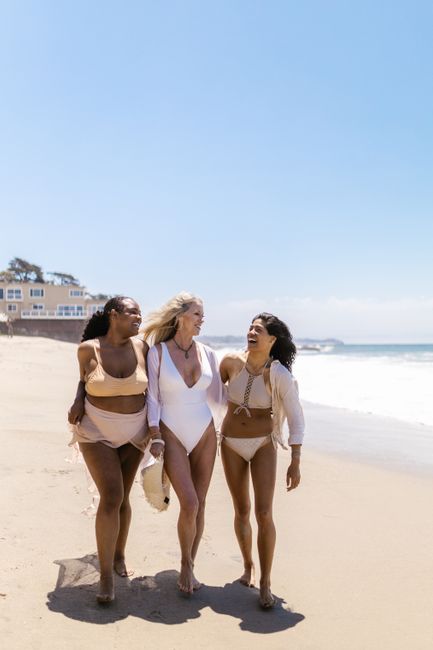 Three women in bathing suits walk and laugh on a sunny beach, enjoying summer.