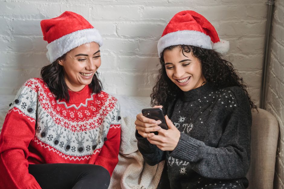 High angle of cheerful ethnic female friends wearing Santa hats laughing together while sharing mobile phone