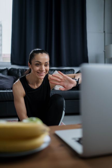 Woman in black fitness attire waving to a video call from home, smiling.