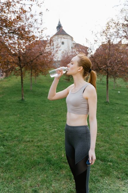 Fit woman drinking bottled water in an outdoor park setting, staying hydrated during workout.
